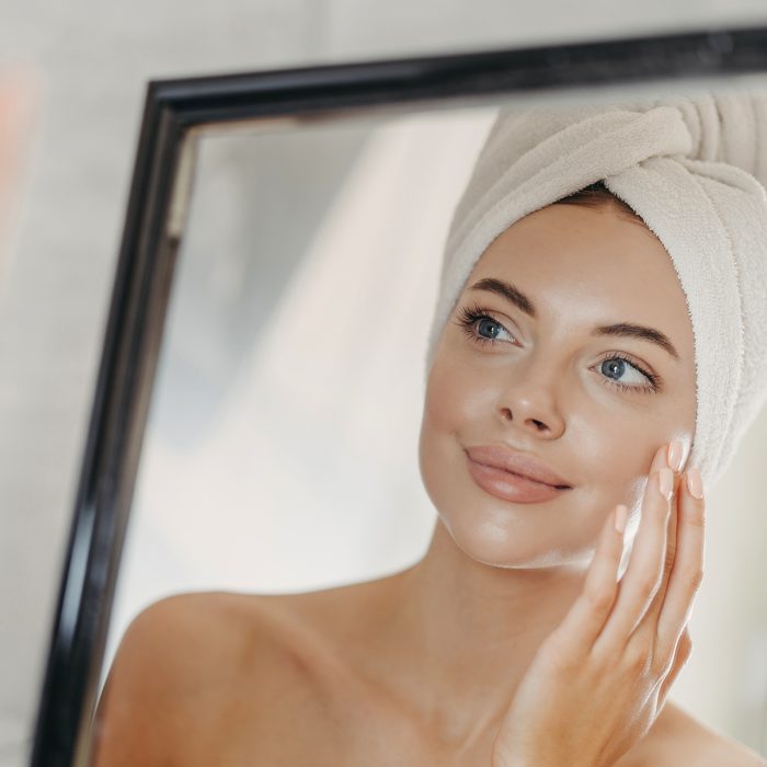 Young naked European woman touches soft skin on face, looks at herself in bathroom mirror, wears wrapped white towel on head after taking bath, poses bare shoulders. Beauty and skin care concept